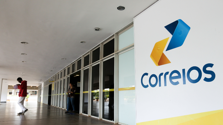 Entrada de agência dos Correios com fachada branca, logo colorida em seta e pessoa caminhando no corredor.