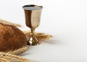 Um cálice dourado com vinho tinto posicionado ao lado de um pão caseiro integral e ramos de trigo seco, sobre um fundo branco limpo.