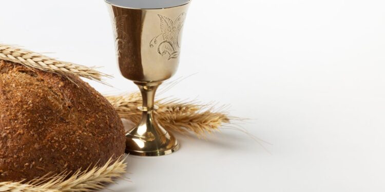 Um cálice dourado com vinho tinto posicionado ao lado de um pão caseiro integral e ramos de trigo seco, sobre um fundo branco limpo.