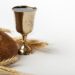 Um cálice dourado com vinho tinto posicionado ao lado de um pão caseiro integral e ramos de trigo seco, sobre um fundo branco limpo.