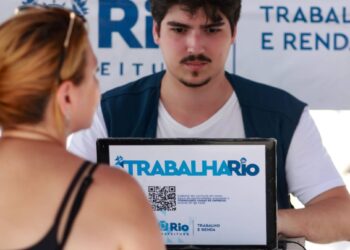 Close-up das mãos de um funcionário da Prefeitura do Rio de Janeiro, vestindo um colete azul escuro com o logo da SMTE, preenchendo dados em uma prancheta durante o atendimento móvel do programa Trabalha Rio. Ao fundo, uma placa branca e azul do programa e o braço de um candidato.