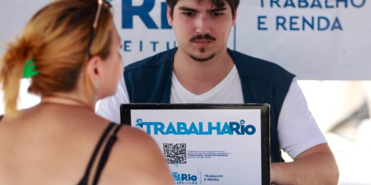 Close-up das mãos de um funcionário da Prefeitura do Rio de Janeiro, vestindo um colete azul escuro com o logo da SMTE, preenchendo dados em uma prancheta durante o atendimento móvel do programa Trabalha Rio. Ao fundo, uma placa branca e azul do programa e o braço de um candidato.