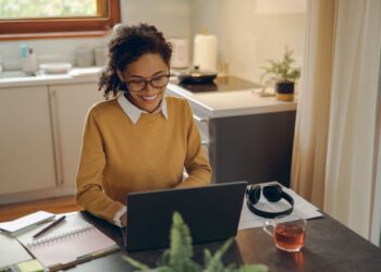 Mulher negra sorrindo enquanto trabalha com um notebook em sua mesa de casa, em um ambiente de cozinha acolhedor e bem iluminado.