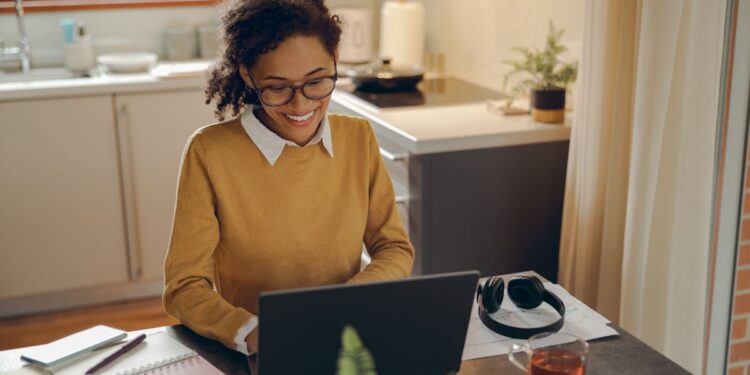 Mulher negra sorrindo enquanto trabalha com um notebook em sua mesa de casa, em um ambiente de cozinha acolhedor e bem iluminado.