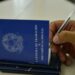 Close de uma mão masculina segurando uma caneta sobre uma Carteira de Trabalho e Previdência Social (CTPS) azul aberta. A mão está prestes a assinar ou preencher o documento sobre uma mesa branca.