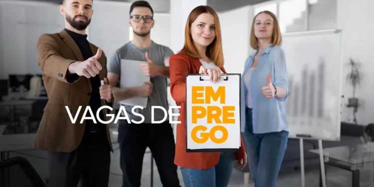 Foto em plano médio de uma equipe de quatro profissionais em pé num escritório moderno. No centro, uma mulher de casaco vermelho segura uma prancheta com o texto laranja: "EM PREGO". Atrás dela, o texto branco flutuante diz: "VAGAS DE". Os outros três profissionais (dois homens e uma mulher) sorriem e fazem sinal de "joia" com o polegar. Fundo claro e desfocado de ambiente corporativo com cadeiras e lousa branca.