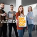 Foto em plano médio de uma equipe de quatro profissionais em pé num escritório moderno. No centro, uma mulher de casaco vermelho segura uma prancheta com o texto laranja: "EM PREGO". Atrás dela, o texto branco flutuante diz: "VAGAS DE". Os outros três profissionais (dois homens e uma mulher) sorriem e fazem sinal de "joia" com o polegar. Fundo claro e desfocado de ambiente corporativo com cadeiras e lousa branca.