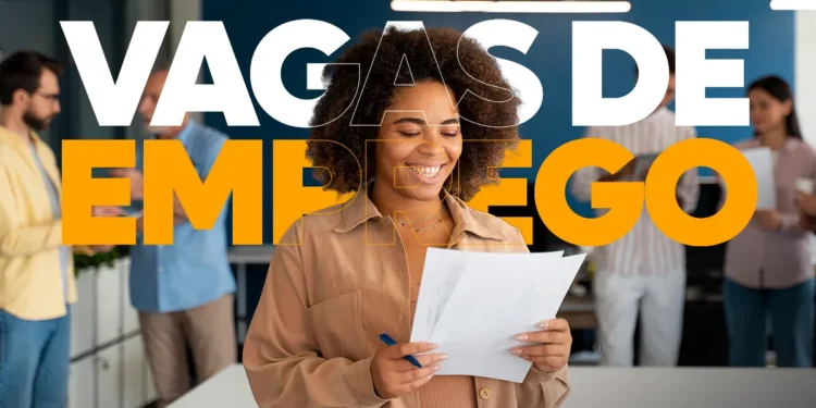 Close-up de uma mulher negra sorridente com cabelos cacheados, vestindo uma camisa marrom-clara, segurando um currículo em papel e uma caneta azul. Sobreposta a ela, há um texto grande em branco e laranja que diz "VAGAS DE EMPREGO". Ela está em um escritório moderno, com outros colegas ao fundo, desfocados.