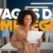 Imagem gráfica com o texto em destaque "VAGAS DE EMPREGO" (em branco e amarelo). Uma mulher sorridente com cabelo afro e vestindo bege lê documentos em primeiro plano, com um grupo borrado ao fundo em um escritório moderno.